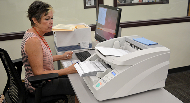 An election worker sits in front of a computer screen and a large scanner stacked with ballots