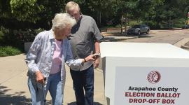 Older female voter inserts ballot into Arapahoe County ballot box while male looks on
